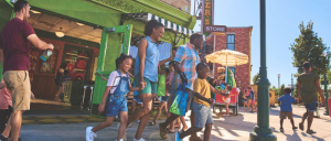 A family walks together outside a colorful store, surrounded by other visitors enjoying a sunny day at a lively outdoor setting.