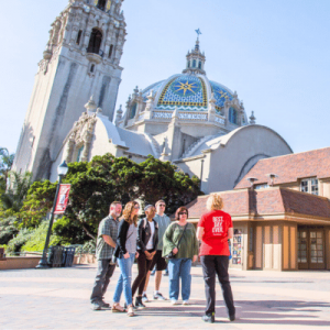 A tour guide in a red shirt speaks to a small group of visitors in front of a grand building with a dome and intricate architecture.